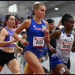 A side-profile action shot of distance runner Peninah Mutisya in a blue and white Hampton University uniform (Bib 182), mid-stride on a bright blue indoor track.