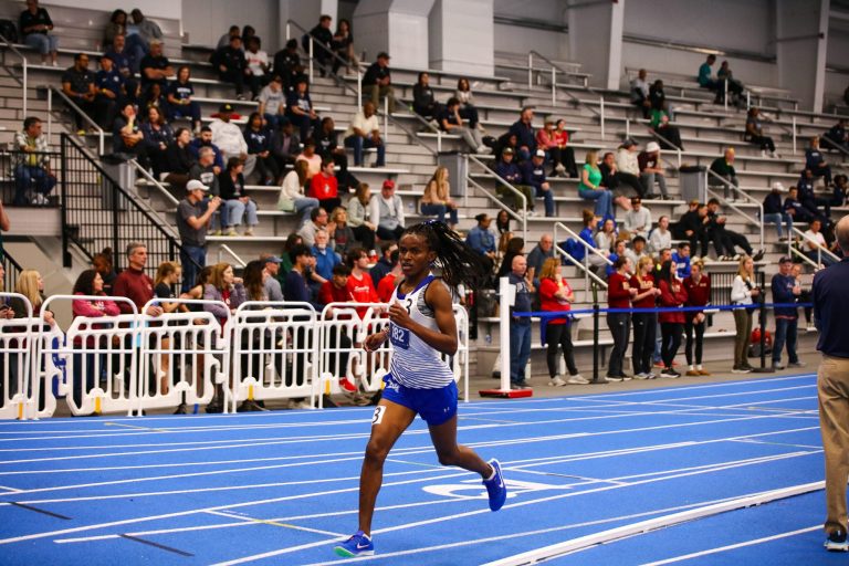 Distance runner Peninah Mutisya of Hampton University wearing a blue and white singlet and bib 182, running on a bright blue indoor track during the 2025 CAA Championships.