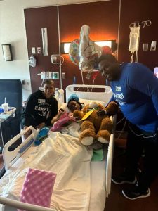 Peninah Mutisya lying in a hospital bed at a U.S. hospital, surrounded by friends visiting her during recovery from a femur fracture
