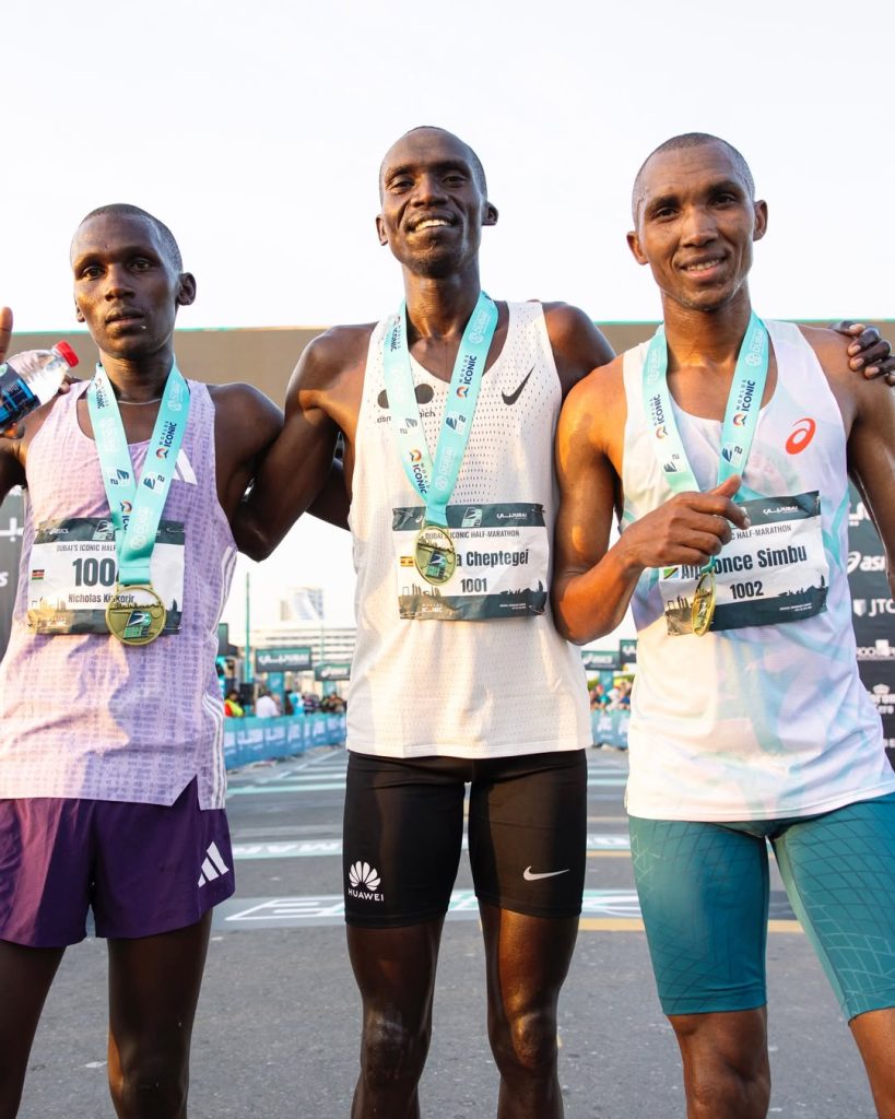 Podium shot of Joshua Cheptegei, Nicholas Kipkorir, and Alphonce Simbu celebrating their top-three finish at the 2026 Burj2Burj Half Marathon.